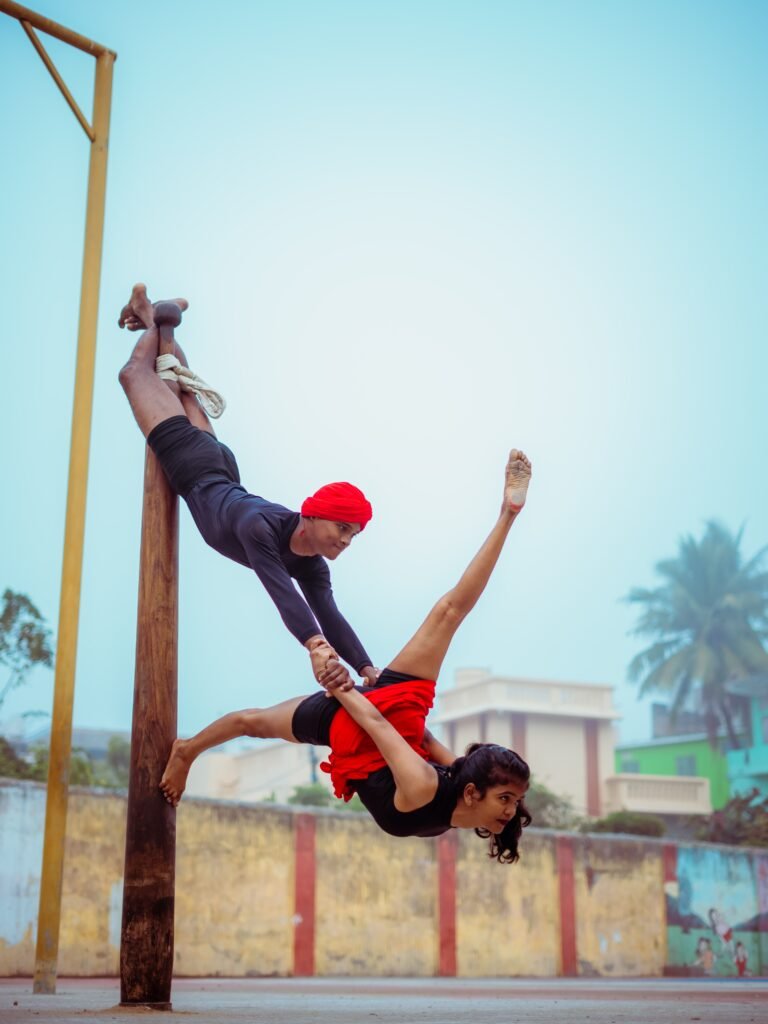 Two Mallakhamb performers balance on a wooden pole one gripping it with his legs, the other suspended horizontally, holding hands.
