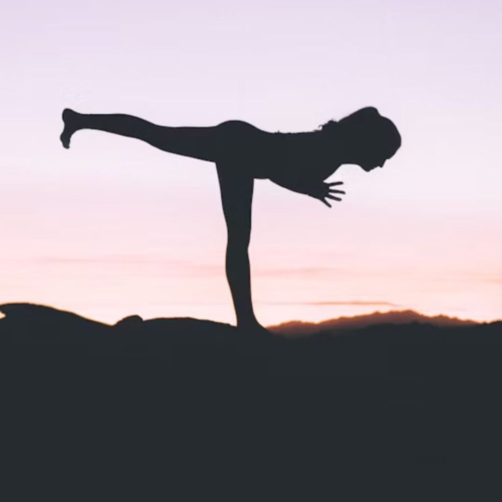 Silhouette of a person balanced on one leg in a yoga pose, against a purple and pink sunset sky.